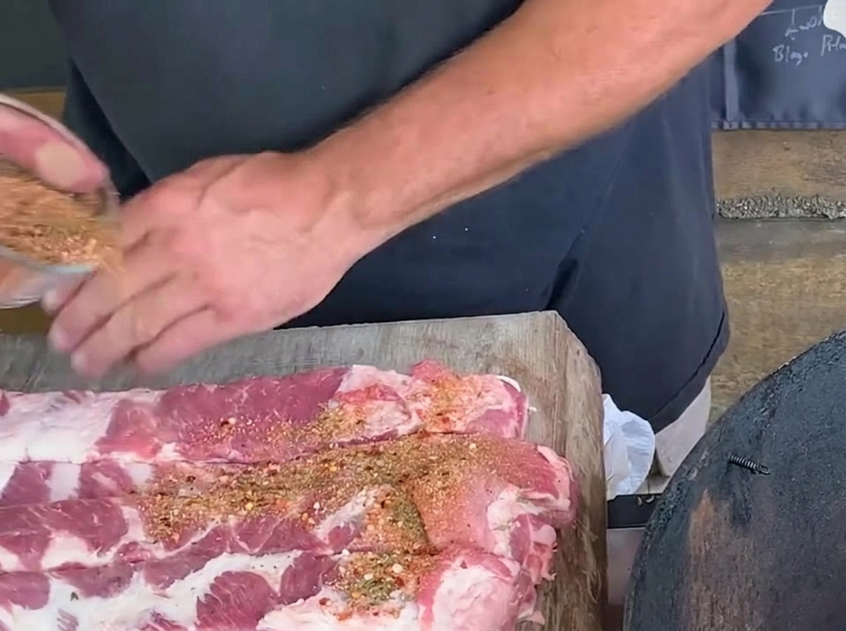 Eine Person streut eine Gewürzmischung aus einer Glasschale auf lange geschnittene rohe Schweinerippchen auf einem rustikalen Holzbrett.
