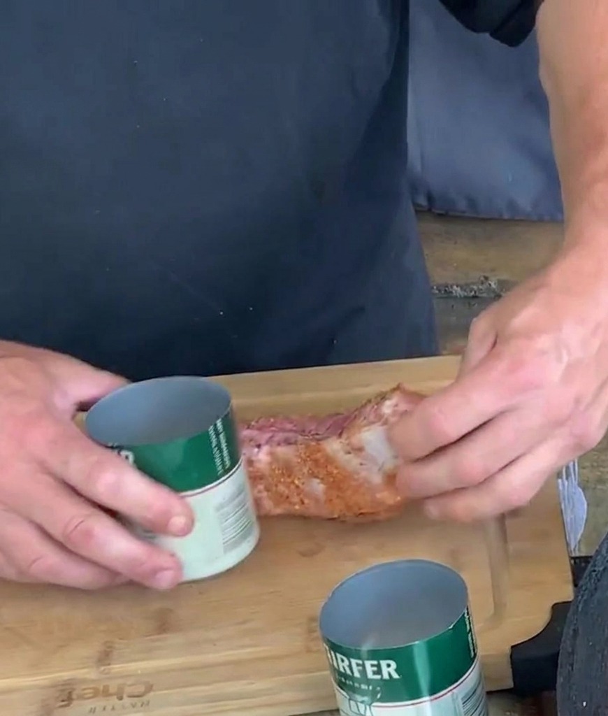 Hände formen einen gewürzten Schweinerippchen-Streifen, indem sie ihn auf einem Schneidebrett um eine offene grün-weiße Aluminiumdose wickeln.
