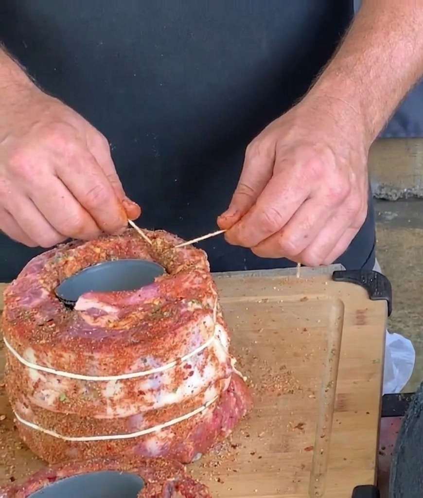 Hände verwenden weißes Küchengarn und einen Zahnstocher, um eine große zylindrische Rolle aus gewürzten Schweinerippchen um eine Dose festzubinden.