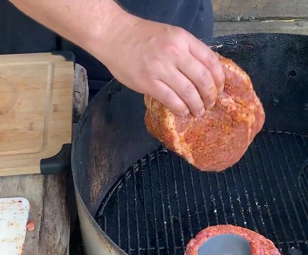 Eine Hand legt einen festgebundenen, gewürzten Rippchen-Becher zum langsamen Garen auf einen schwarzen Holzkohlegrillrost.