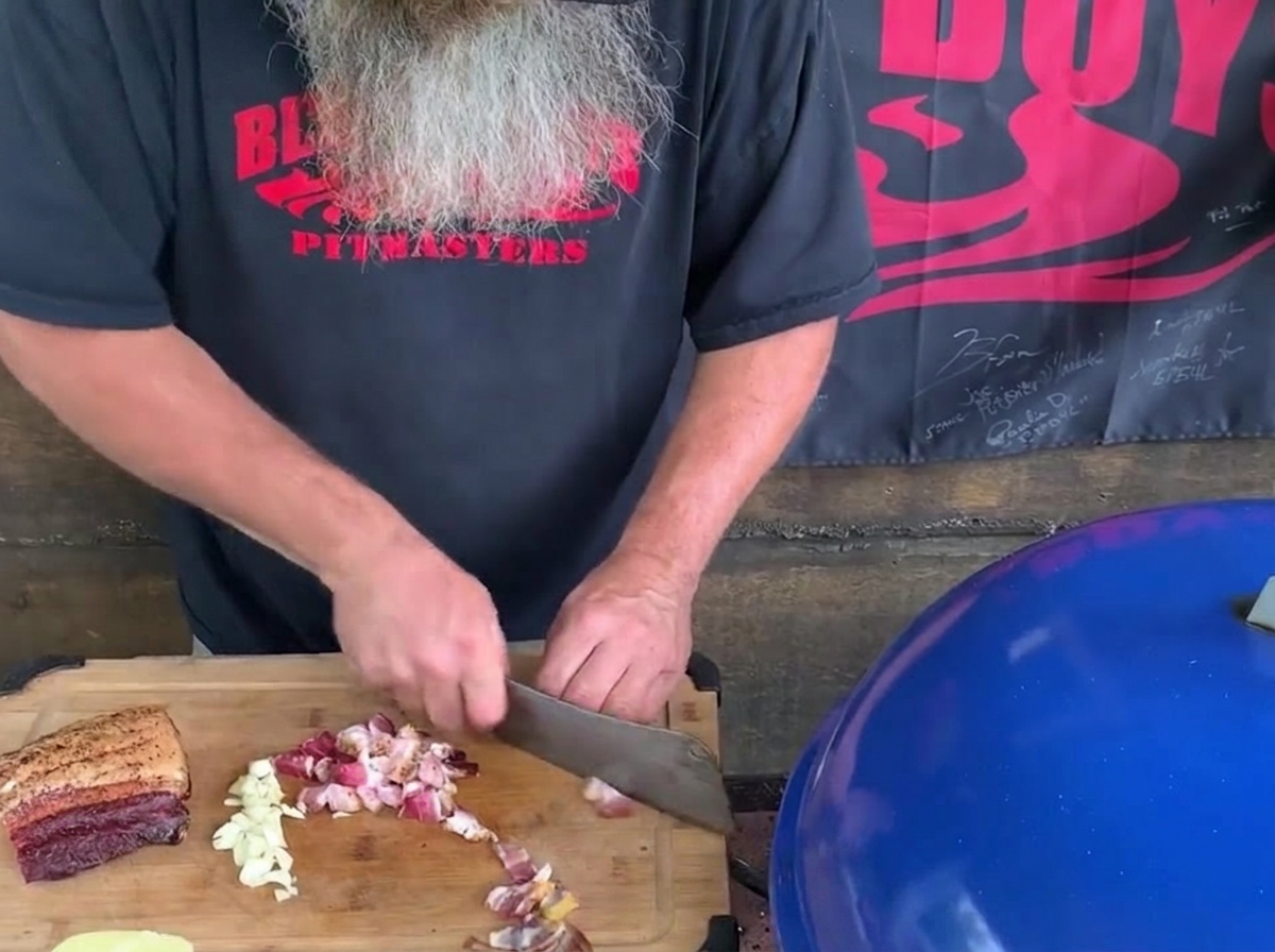 Ein man mit langem grauem Bart hackt Speckwürfel und Knoblauchzehen mit einem großen Küchenbeil auf einem Holzschneidebrett neben einem blauen Grill.