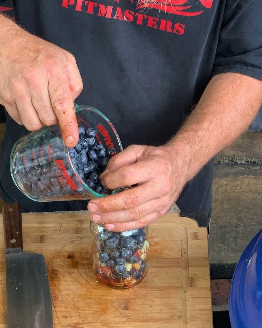 Eine Nahaufnahme von Händen, die frische Blaubeeren aus einem Pyrex-Messbecher in ein Einmachglas gießen, das bereits flüssige und feste Saucenzutaten enthält.