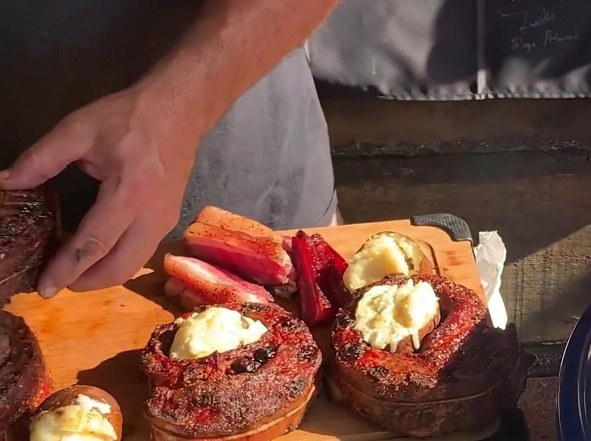 Eine Hand legt eine heiße, mit Butter bestrichene Röstkartoffel in die hohle Mitte eines gegarten, runden Rippchen-Bechers auf einem Holzschneidebrett.