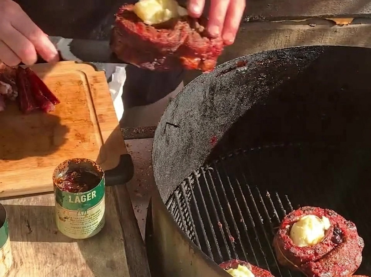 Ein gefüllter Rippchen-Becher wird neben einem weiteren Fleischbecher und dicken Speckscheiben auf einen Holzkohlegrill gelegt.