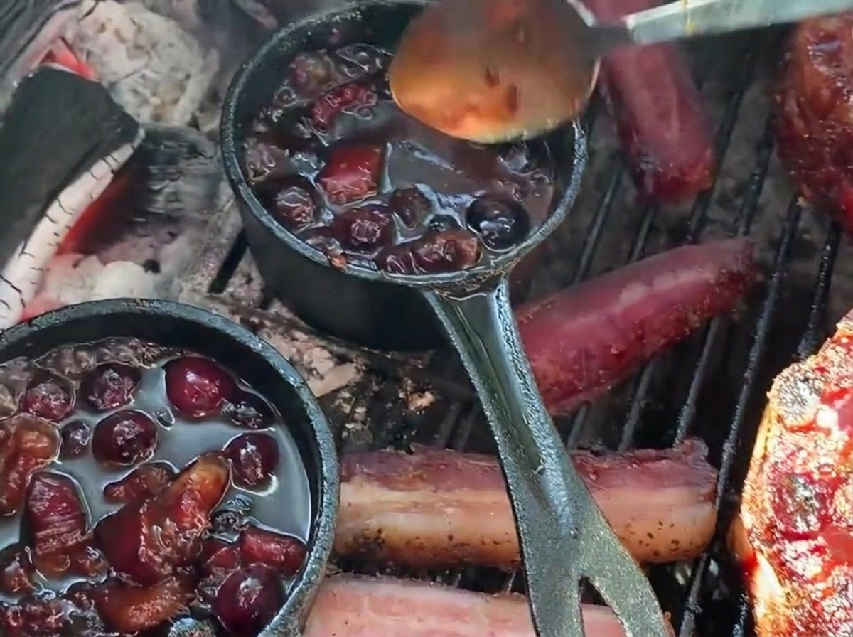 Reichhaltige Blaubeer-Barbecue-Sauce wird mit einem Löffel aus einem kleinen Gusseisentopf über einen gefüllten Rippchen-Becher auf dem Grill gegossen.