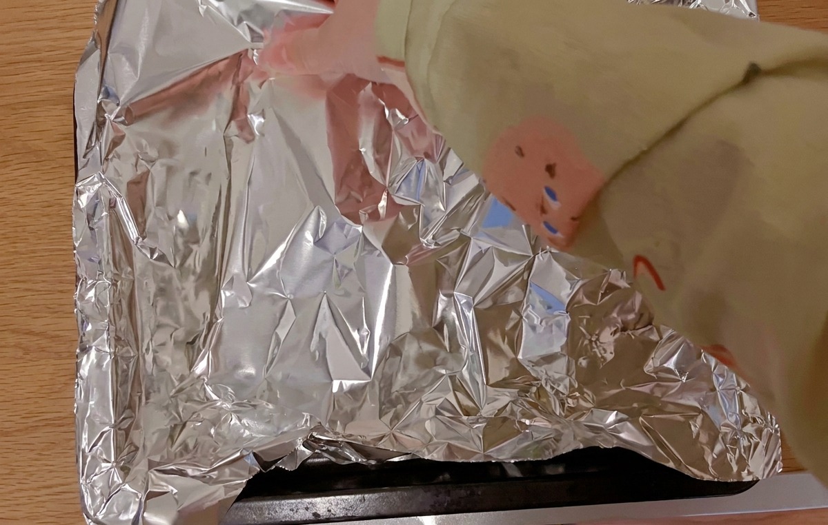 Hände decken marinierte Schweinerippchen auf einem schwarzen Backblech mit einem Stück glänzender Alufolie ab.