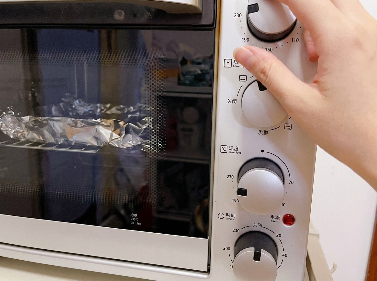 Nahaufnahme einer Hand, die den Temperaturregler eines weißen Elektro-Miniofens auf 230°C einstellt, im Inneren sind in Folie gewickelte Speisen auf dem Rost zu sehen.