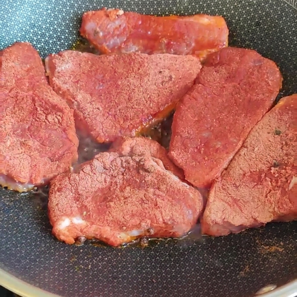 Dicke Schweinenackenscheiben, die mit einer roten Gewürzmischung bedeckt sind, braten in einer heißen Pfanne auf dem Herd.