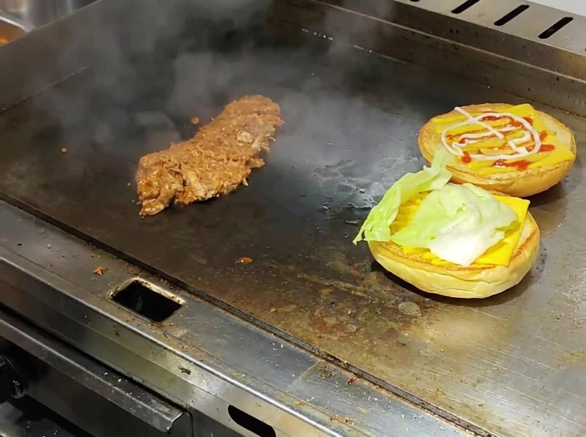 Angetoastete Burger-Brötchen, belegt mit Käse und Salat, neben heißem Pulled Pork auf einer dampfenden Grillplatte.
