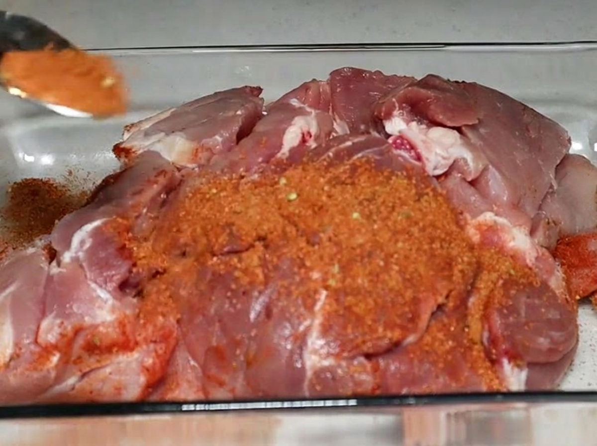 Ein Löffel rötliche Gewürzmischung wird über ein großes Stück rohe Schweineschulter gestreut, das in einer Glasauflaufform liegt.