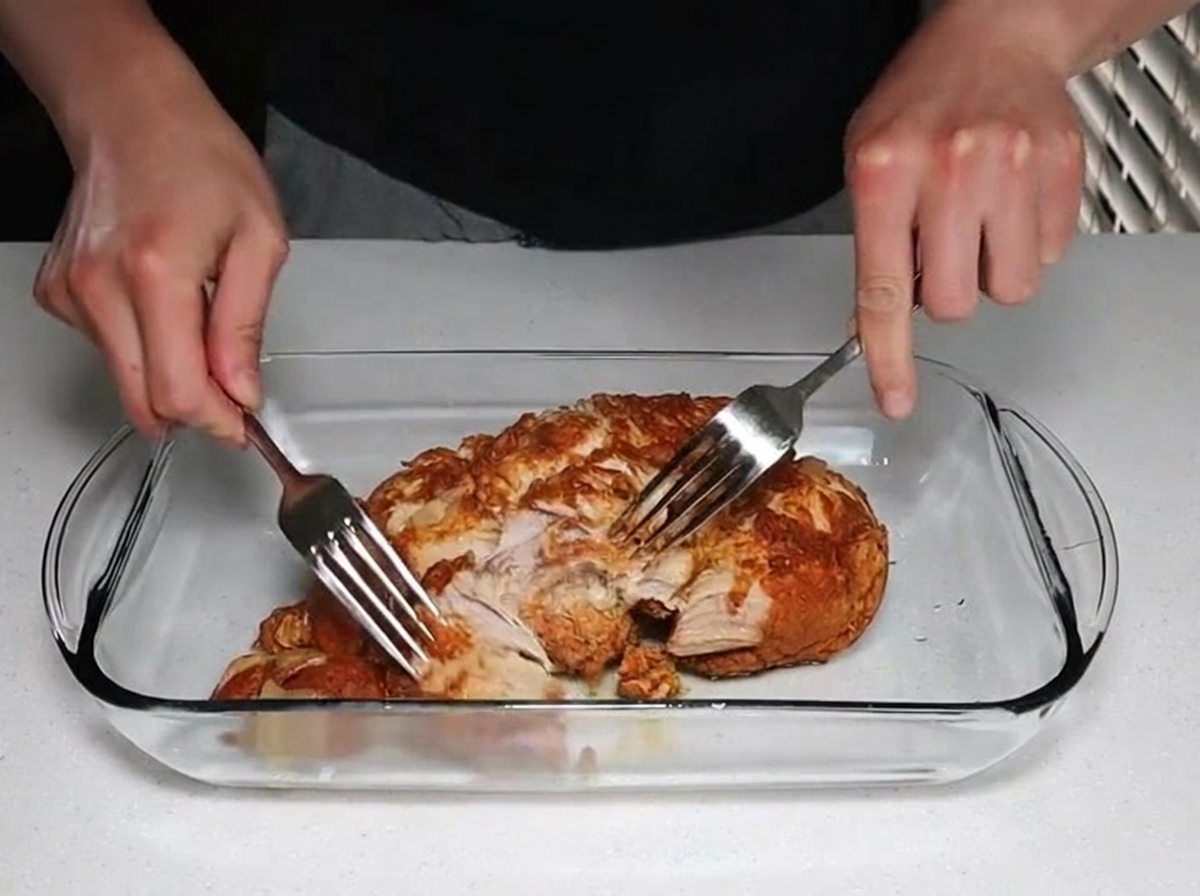 Zwei Hände zerzupfen ein großes Stück zartes, gegartes Schweinefleisch mit zwei Edelstahlgabeln in einer klaren Glasauflaufform.