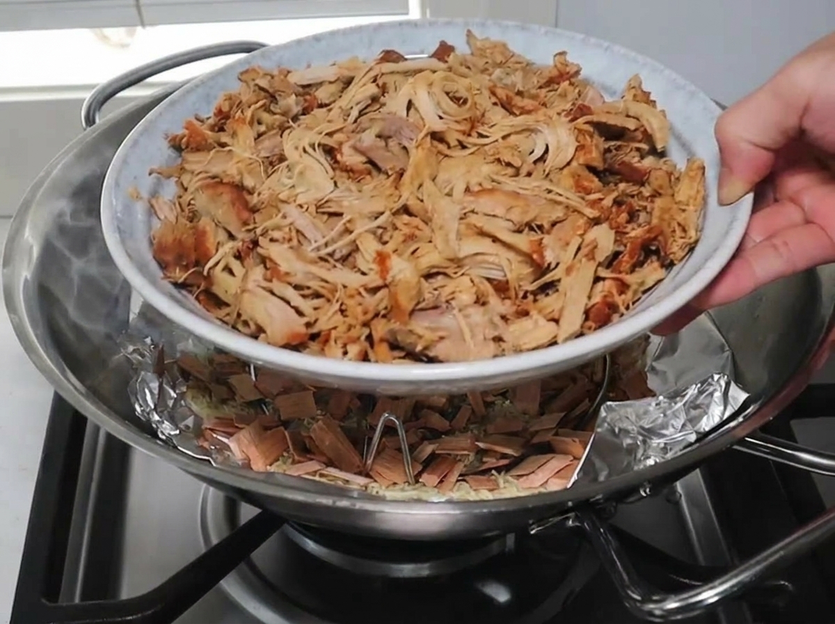 Eine Hand stellt einen weißen Teller mit aufgehäuftem Pulled Pork auf einen Dämpfeinsatz im Wok über den rauchenden Holzchips.