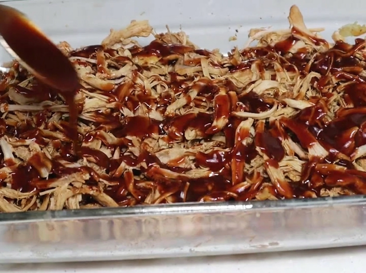 Ein Löffel dicke, dunkle BBQ-Sauce wird über eine Schicht aus geräuchertem Pulled Pork in einer klaren Glasauflaufform gegossen.