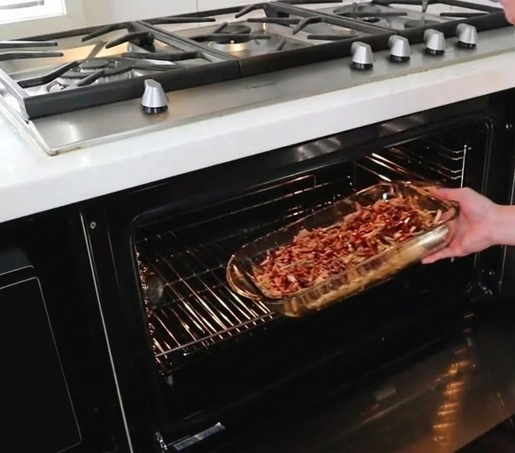 Eine Person schiebt eine Glasauflaufform mit mariniertem Pulled Pork auf die mittlere Schiene eines heißen Backofens.