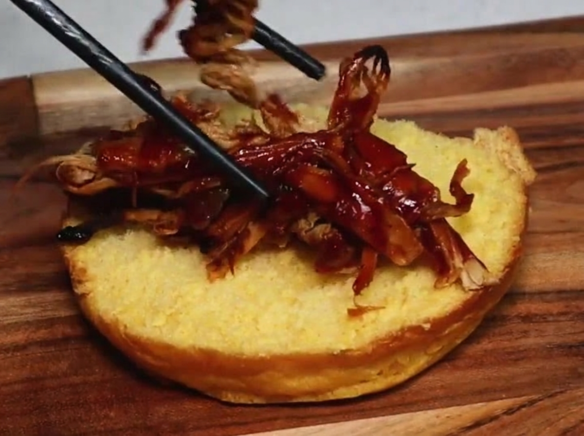 Ein Paar Essstäbchen legt saftiges Pulled Pork mit dunkler BBQ-Sauce auf die goldbraune Unterseite eines getoasteten Burger-Brötchens.