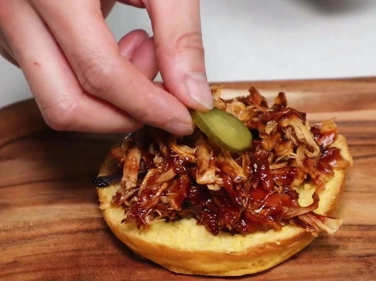 Eine Hand legt eine runde Gewürzgurkenscheibe auf einen Haufen Pulled Pork auf einem getoasteten Burger-Brötchen.