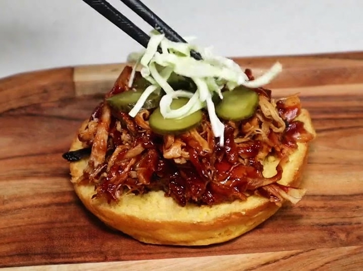 Essstäbchen legen feinen Krautsalat auf die Gurkenscheiben und das Pulled Pork in einem Burger-Brötchen.