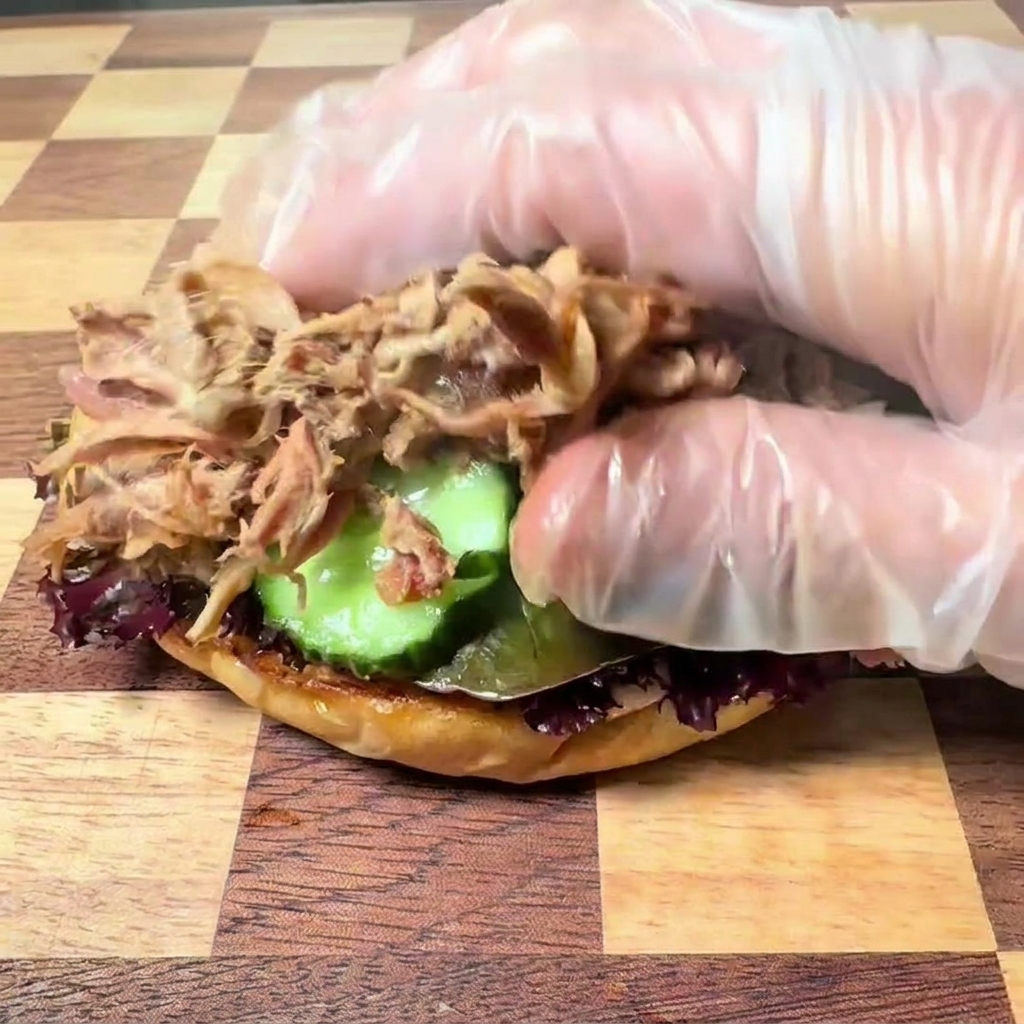 Eine Hand mit transparentem Plastikhandschuh legt zartes, zerzupftes Pulled Pork auf eine Burger-Unterseite mit Salat und Gurkenscheiben.