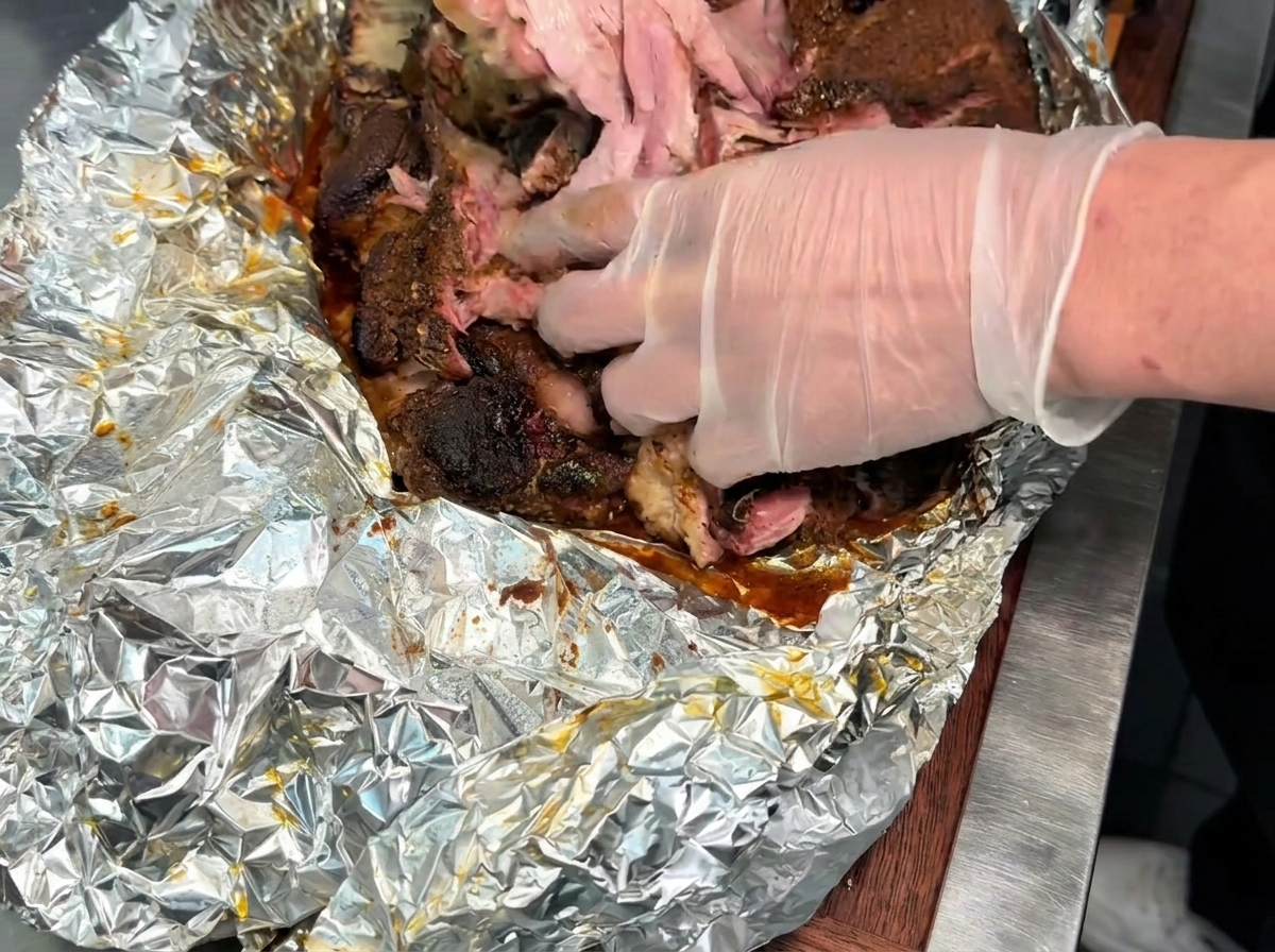 Behandschuhte Hände zerkleinern und zerzupfen ein großes Stück zartes, gegartes Räucherschweinefleisch auf einer mit Folie ausgelegten Oberfläche.