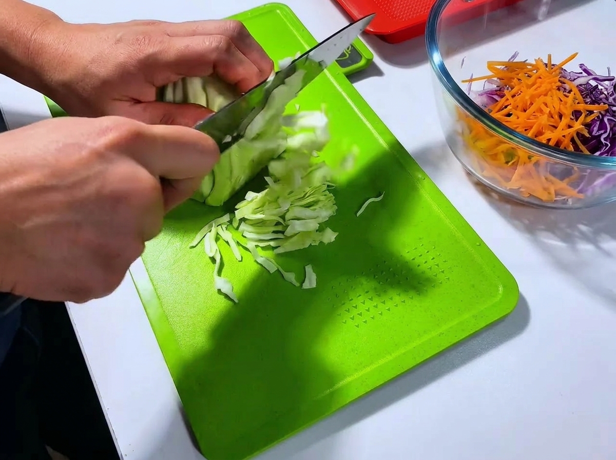 Ein Koch schneidet einen Weißkohl auf einem grünen Schneidebrett mit einem großen Messer in sehr feine Streifen.