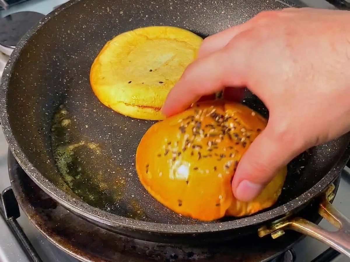 Eine Hand wendet eine goldbraun geröstete Burgerbrötchen-Hälfte in einer Pfanne mit geschmolzener Butter.