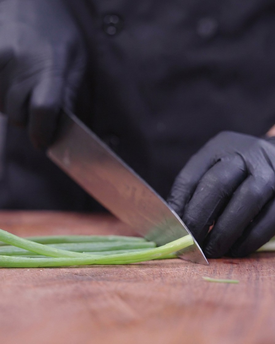 Ein Koch mit schwarzen Handschuhen schneidet frische Frühlingszwiebeln mit einem großen Messer auf einem Holzbrett.