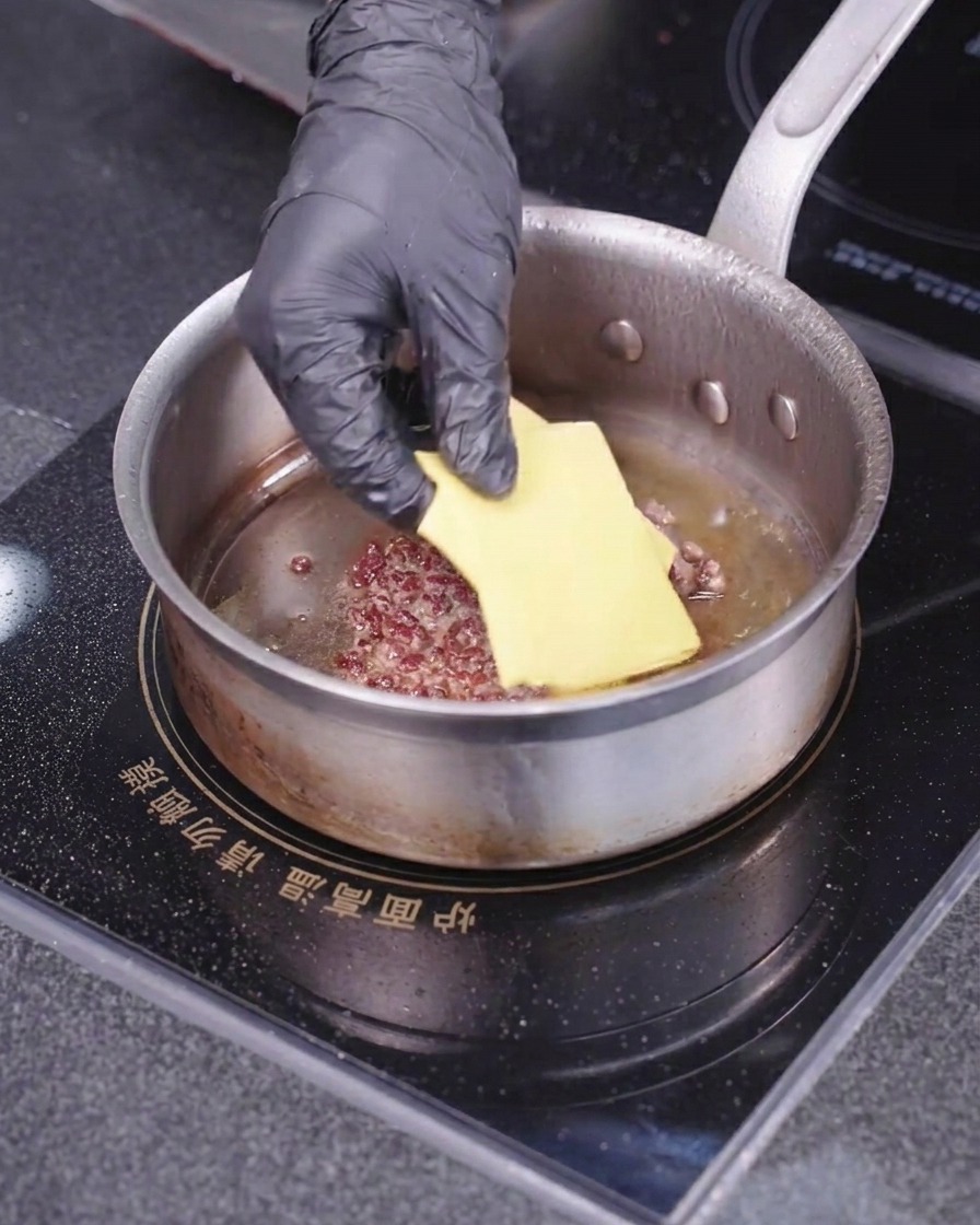 Eine behandschuhte Hand legt eine quadratische Scheibe gelben Käse auf ein bratendes Fleisch-Patty in einer heißen Pfanne.