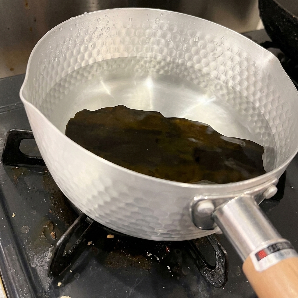 Ein Stück dunkler, getrockneter Seetang liegt am Boden eines gehämmerten Aluminiumtopfs mit klarem, kaltem Wasser.