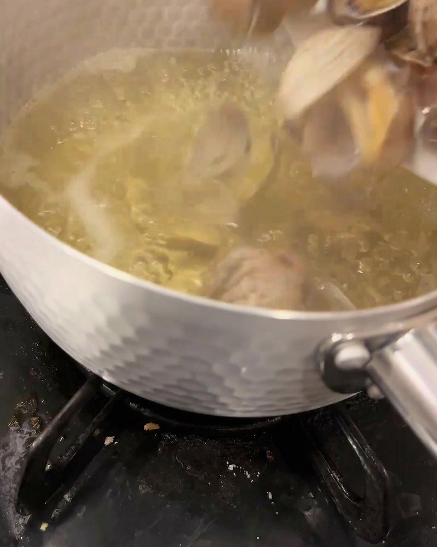 Frische Muscheln werden in eine kochende, klare Dashi-Brühe in einem Metalltopf auf dem Gasherd gegeben.
