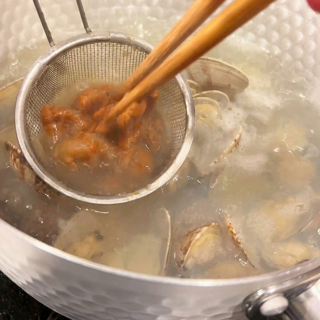 Ein kleines Küchensieb hält braune Misopaste, während Essstäbchen sie in die heiße Muschelsuppe einrühren.