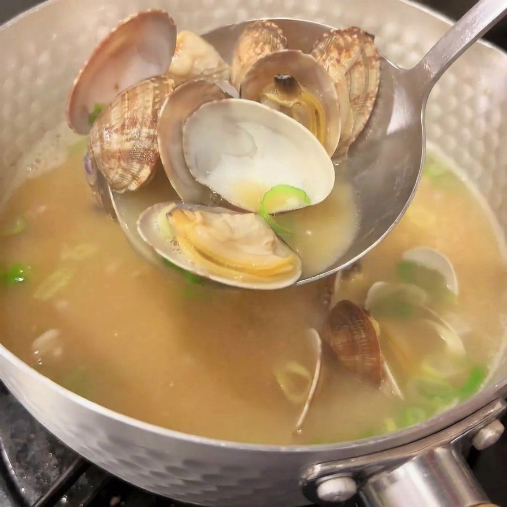 Eine Schöpfkelle hebt geöffnete Muscheln aus einem Topf mit dampfender japanischer Misosuppe, in der frische gehackte Frühlingszwiebeln schwimmen.