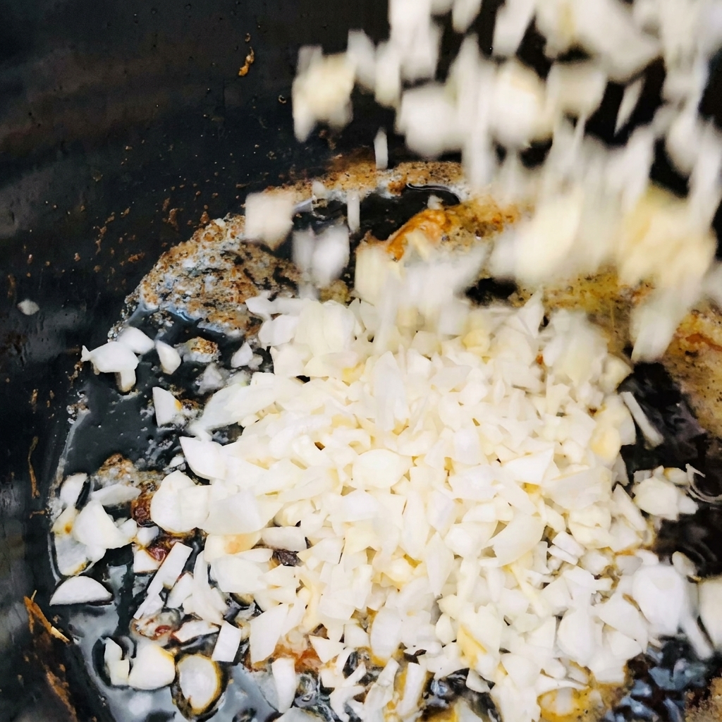 Gewürfelte weiße Zwiebeln und gehackter Knoblauch fallen in einen dunklen Topf mit heißem, schimmerndem Öl.