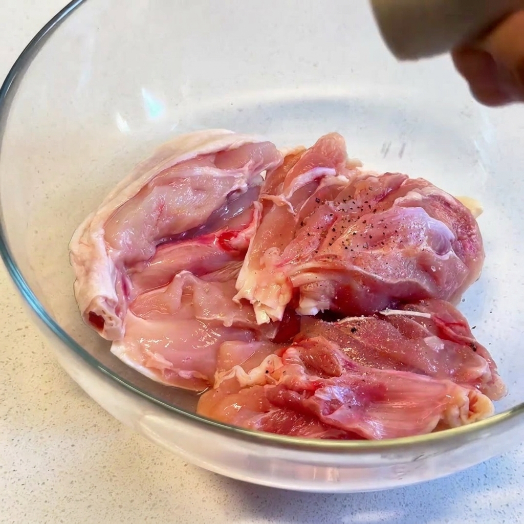 Rohe Hähnchenschenkel in einer Glasschüssel, die mit frisch gemahlenem schwarzen Pfeffer aus einer Mühle gewürzt werden.