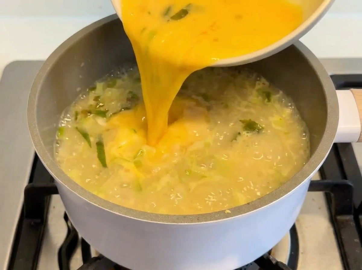 Eine leuchtend gelbe Ei-Zitronen-Mischung wird aus einer weißen Schüssel in einen Topf mit köchelnder, cremiger Suppe gegossen.