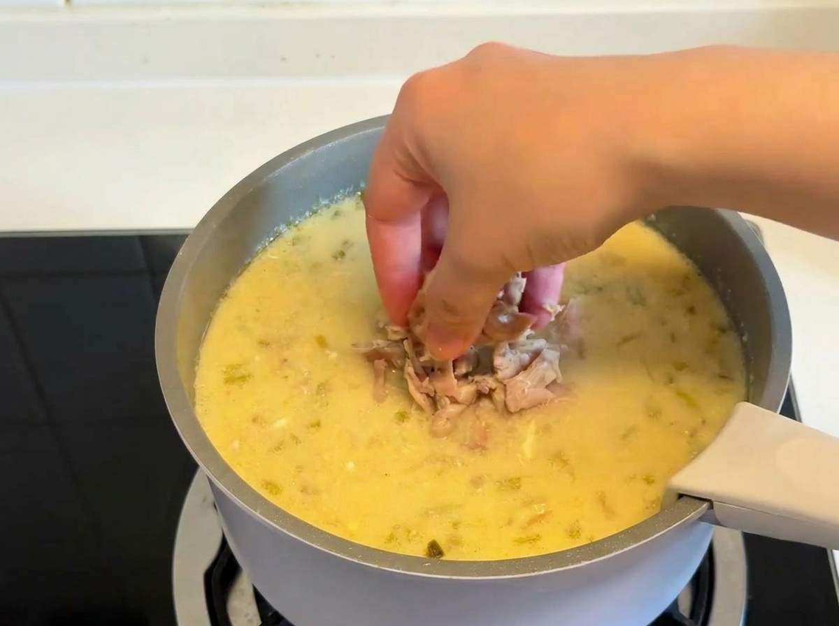 Eine Hand gibt Stücke von gekochtem, gezupftem Hähnchenfleisch in einen Topf mit dicker, cremiger Zitronensuppe.