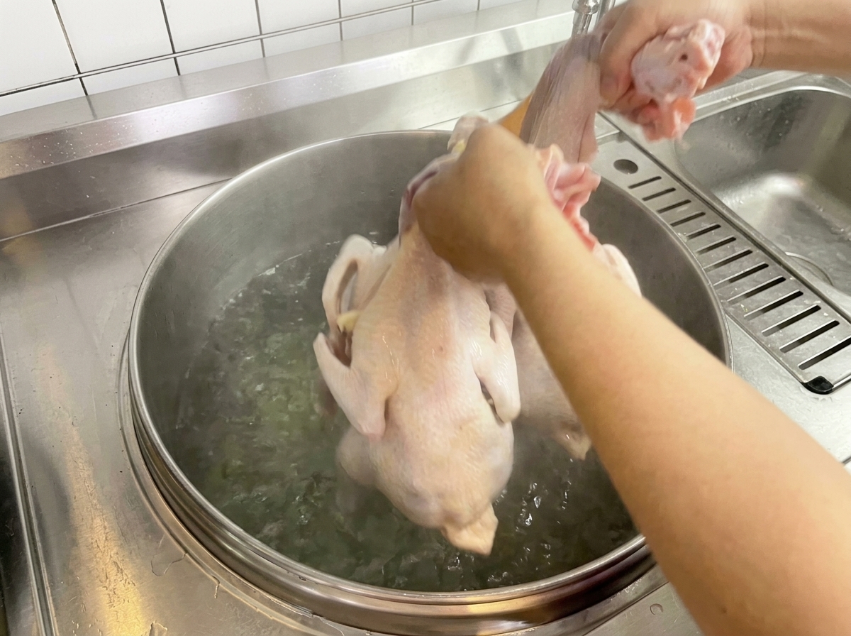 Ein rohes Huhn wird von Hand in einen großen Topf mit kochendem Wasser getaucht.