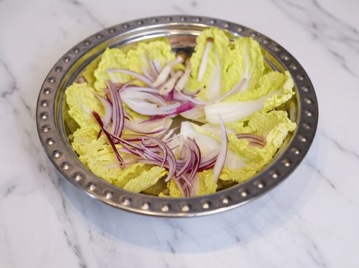 Ein flacher Metalltopf, in dem Kohl, Zwiebelstreifen, Pilze, Fischkuchen und Scheiben von Frühstücksfleisch ordentlich angerichtet sind.