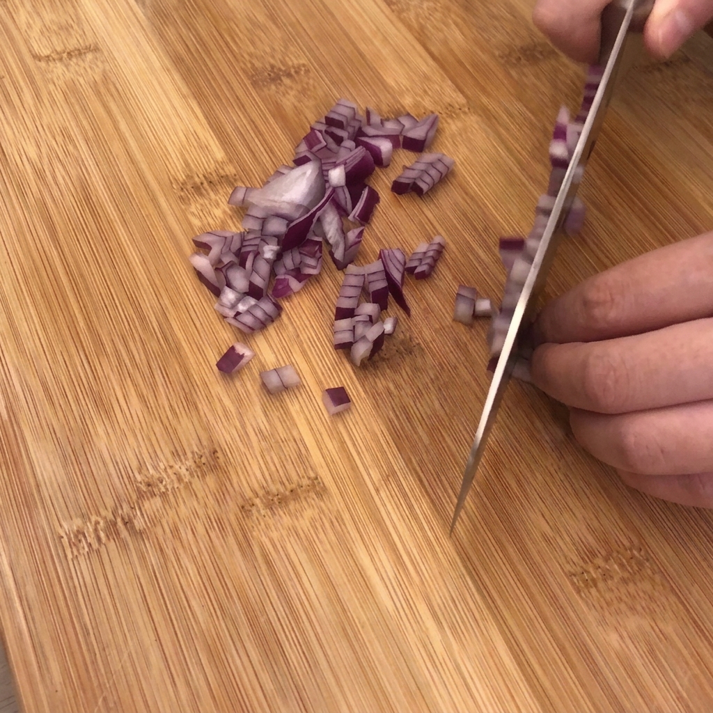 Ein scharfes Küchenmesser schneidet eine rote Zwiebel auf einem Holzschneidebrett in feine Würfel, daneben liegen Knoblauch und Speck.
