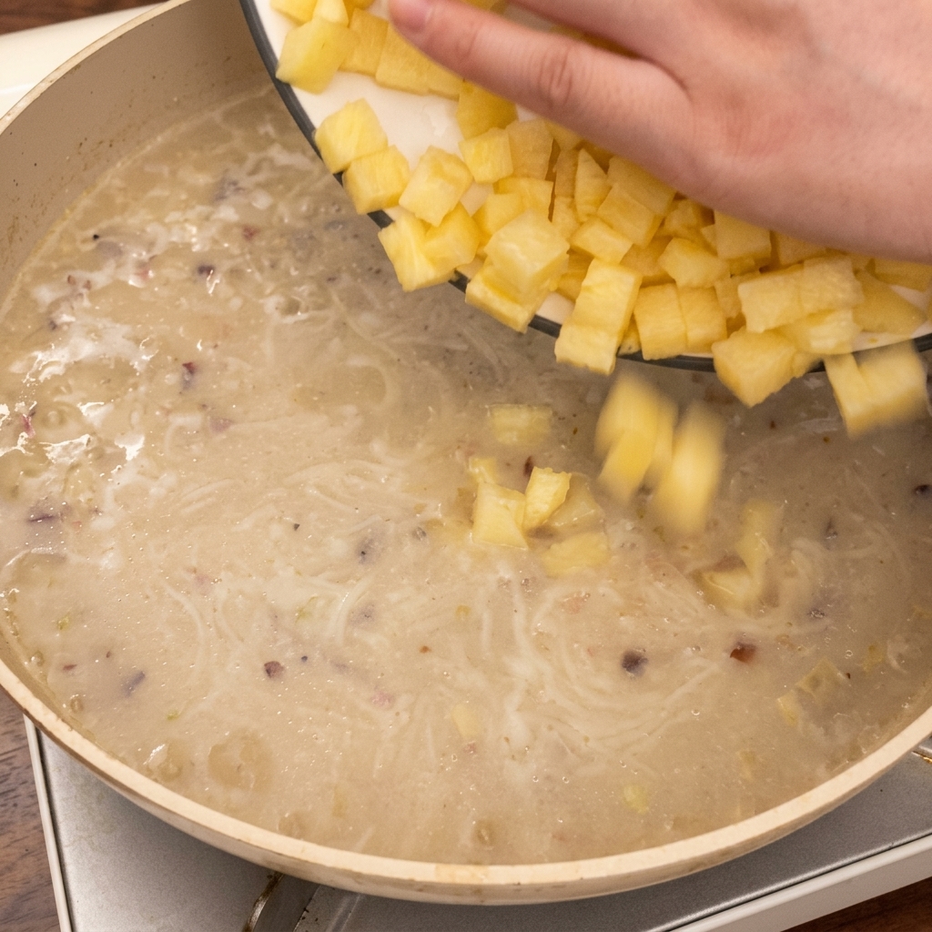 Eine Hand gibt gewürfelte Kartoffeln in die köchelnde Suppe in der Pfanne.