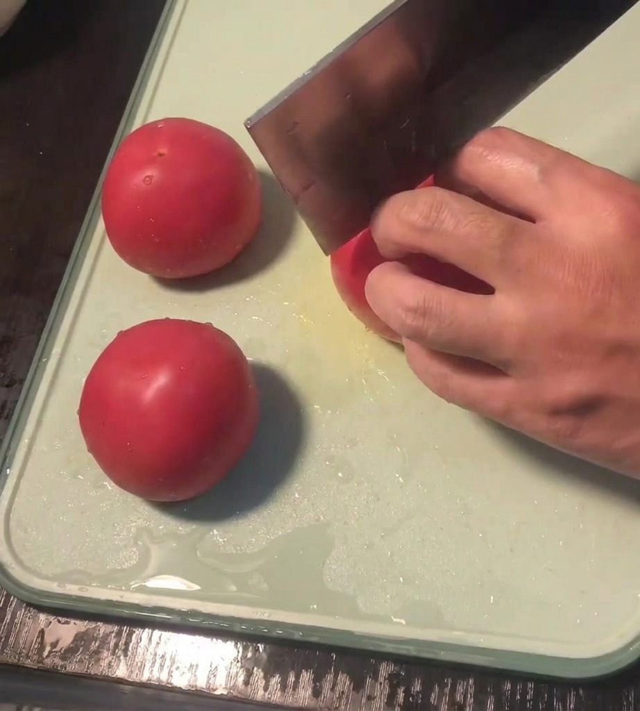 Eine Hand ritzt mit einem Messer vorsichtig ein Kreuz in die Unterseite einer frischen Tomate auf einem Schneidebrett.