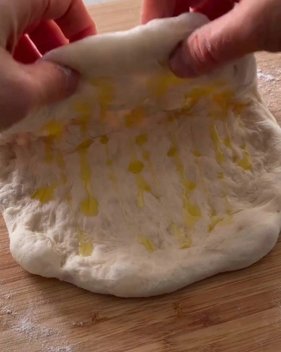Hände falten den geölten Pizzateig auf einer Holzoberfläche in der Mitte zusammen.
