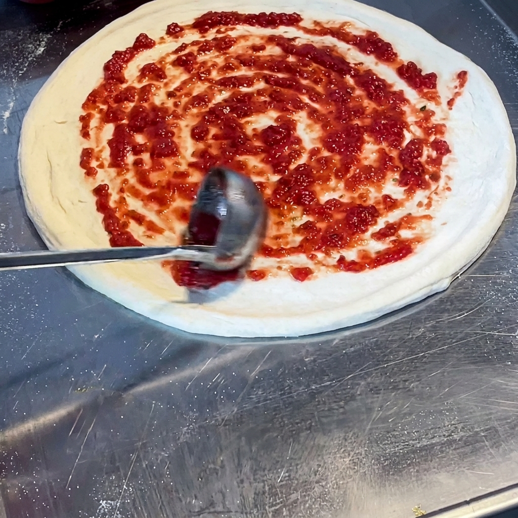 Eine Schöpfkelle verteilt rote Tomatensoße in einer Spiralbewegung auf dem rohen Pizzateig.