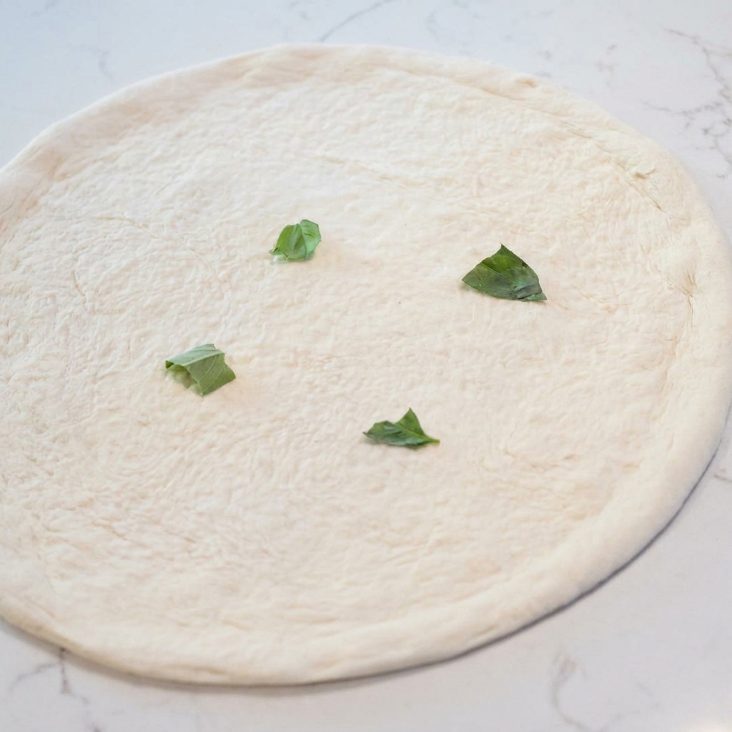 Ein roher, geformter Pizzaboden, belegt mit verteilten frischen grünen Basilikumblättern.