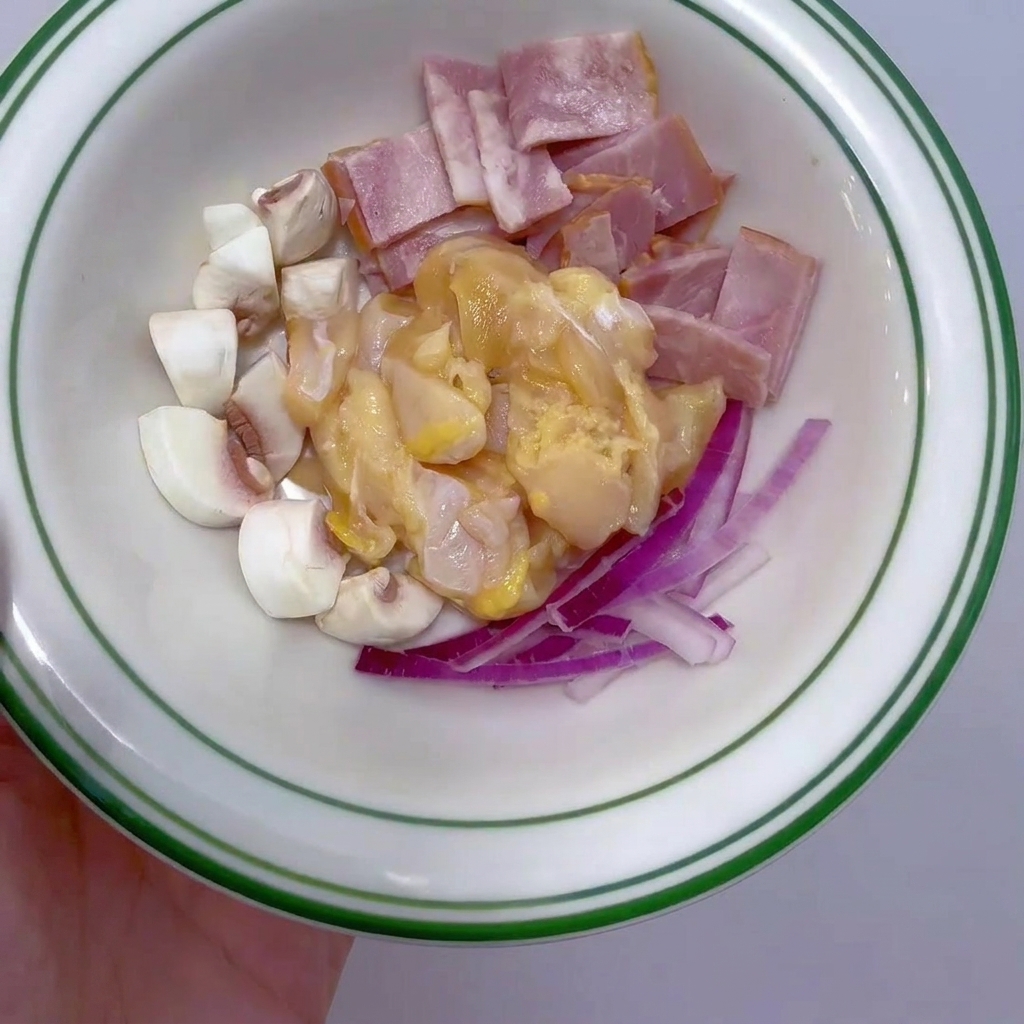 Eine kleine weiße Schüssel mit rohen Hähnchenbrustwürfeln, Speckstreifen, geviertelten weißen Champignons und feinen roten Zwiebelstreifen.