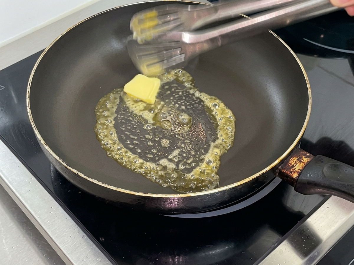 Nahaufnahme eines gelben Butterwürfels, der in einer vorgeheizten schwarzen beschichteten Pfanne schmilzt, im Hintergrund eine Küchenzange.