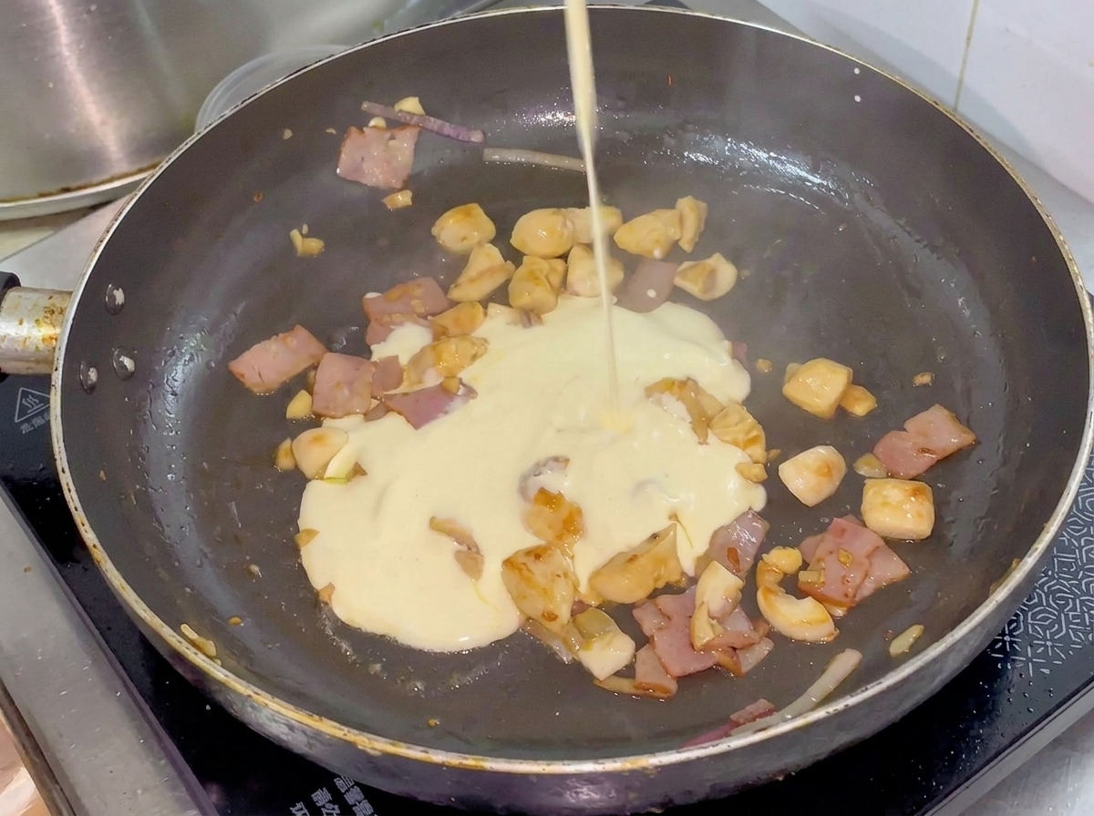 Sahne wird aus einem kleinen Kännchen in eine Pfanne mit gebratenem Hähnchen, Speck und Gemüse gegossen.