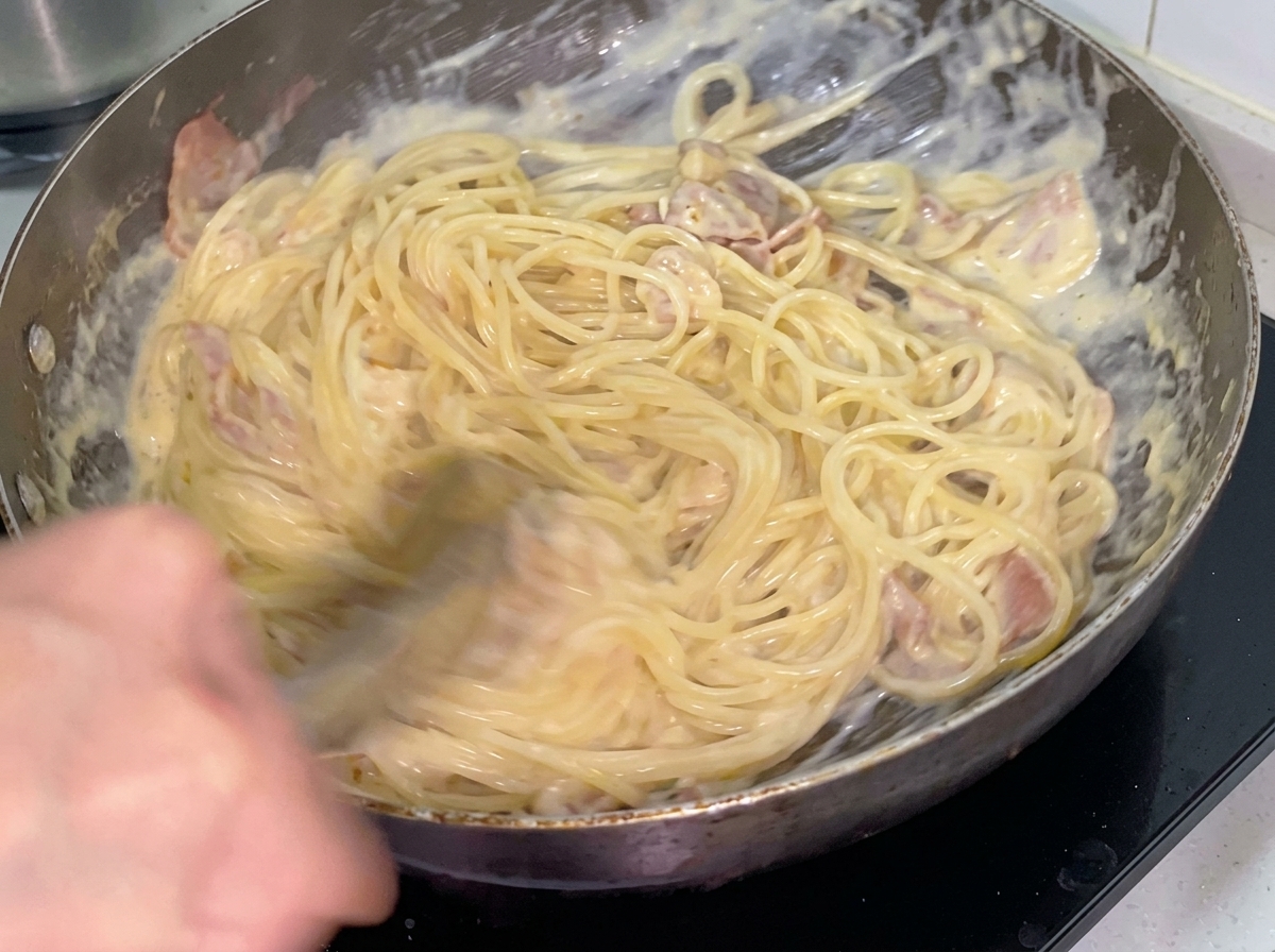 Cremige Spaghetti werden mit einer Metallzange in einer Bratpfanne mit Speck und Hähnchen vermengt.