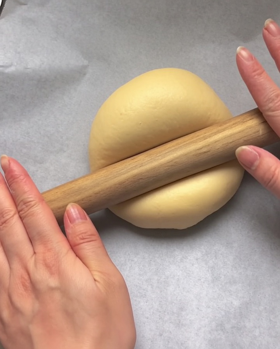 Hände nutzen ein hölzernes Nudelholz, um eine Kugel rohen Teig auf weißem Backpapier kreisförmig auszurollen.