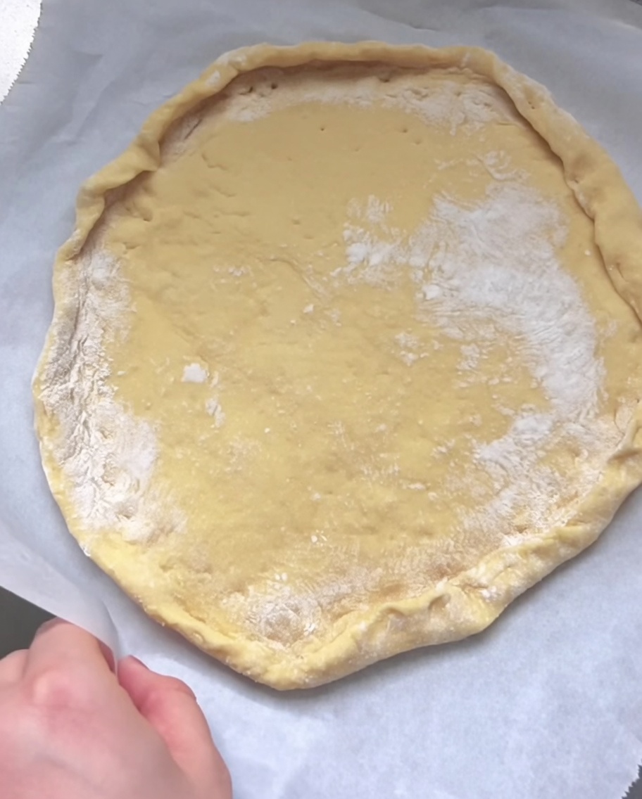 Eine Hand faltet und drückt den Rand des flachen, rohen Teigs, um auf Backpapier einen erhöhten Pizzarand zu formen.