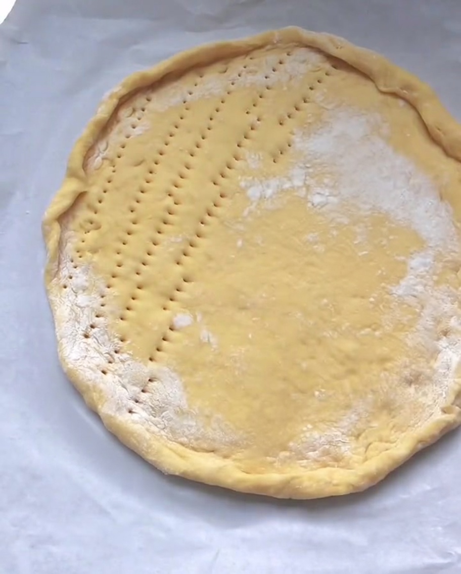 Ein roher, kreisförmiger Pizzaboden auf Backpapier mit kleinen Löchern, die gleichmäßig über die flache Mitte verteilt sind.